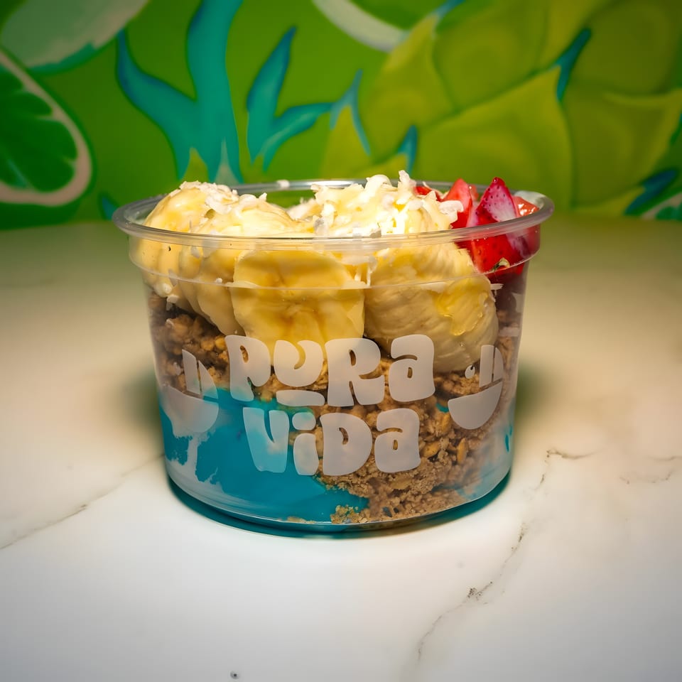 Pura Vida branded cup with blue base, granola, banana slices, shredded coconut, and diced strawberries.