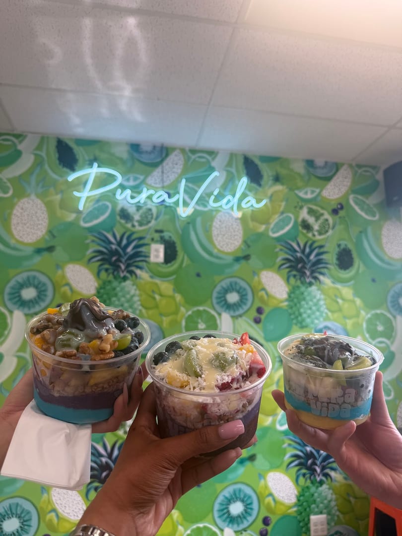 Three hands hold colorful fruit bowls in front of a green Pura Vida wall.