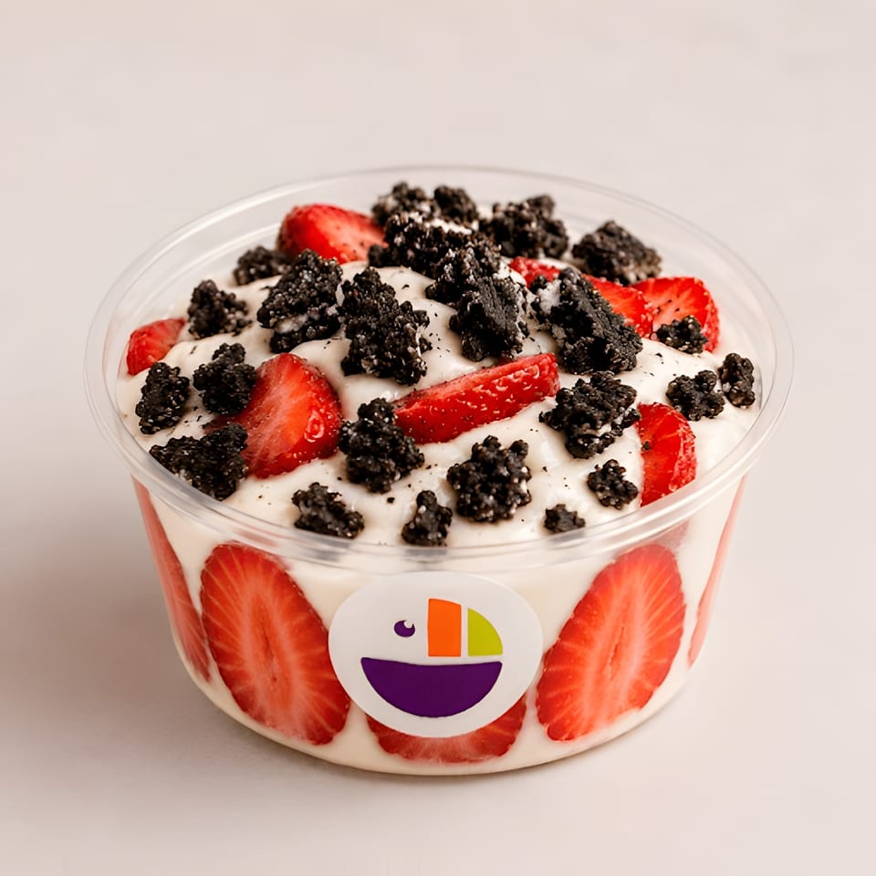 A clear plastic bowl of creamy dessert with sliced strawberries and dark cookie crumbles.